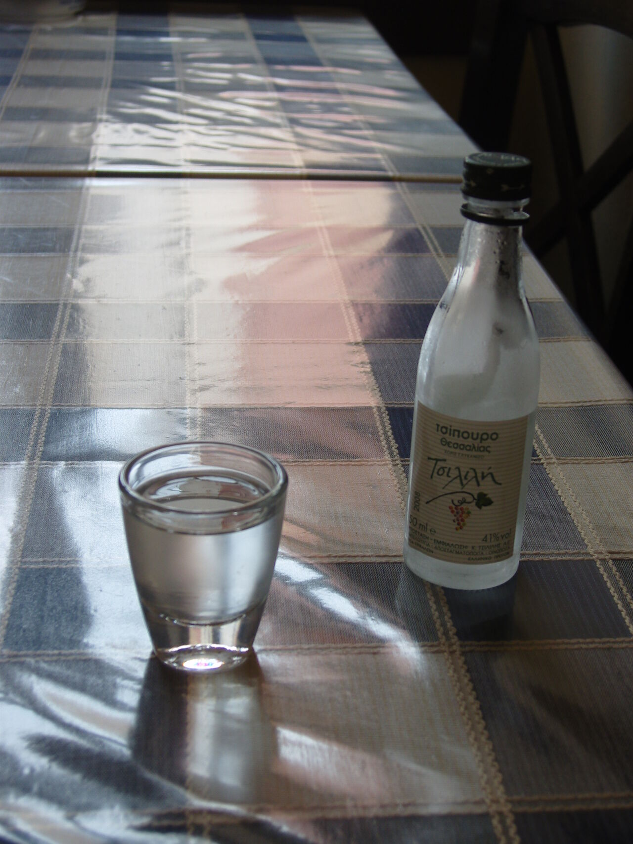 A glass of tsipouro beside its bottle on a checked tablecloth