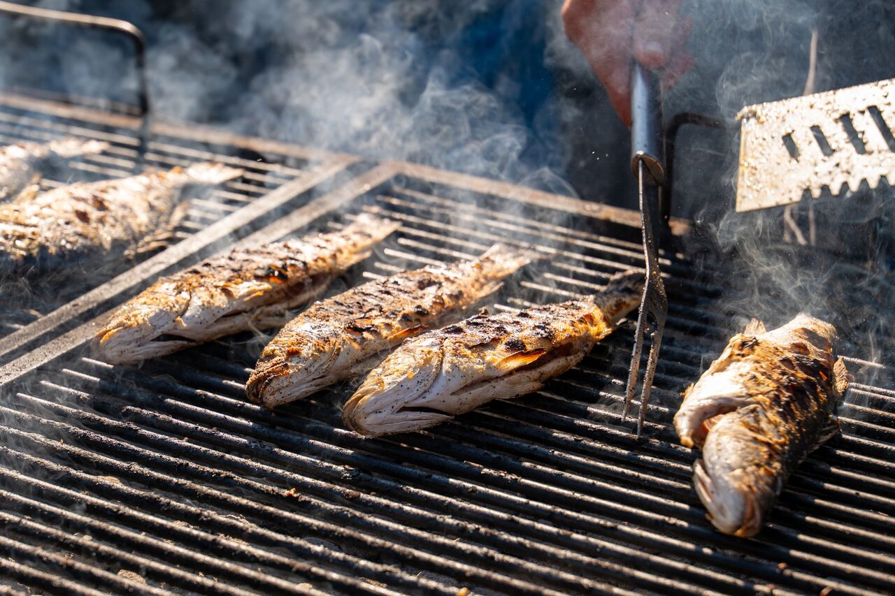 Whole fish on a charcoal grill, smoke rising, tongs turning them