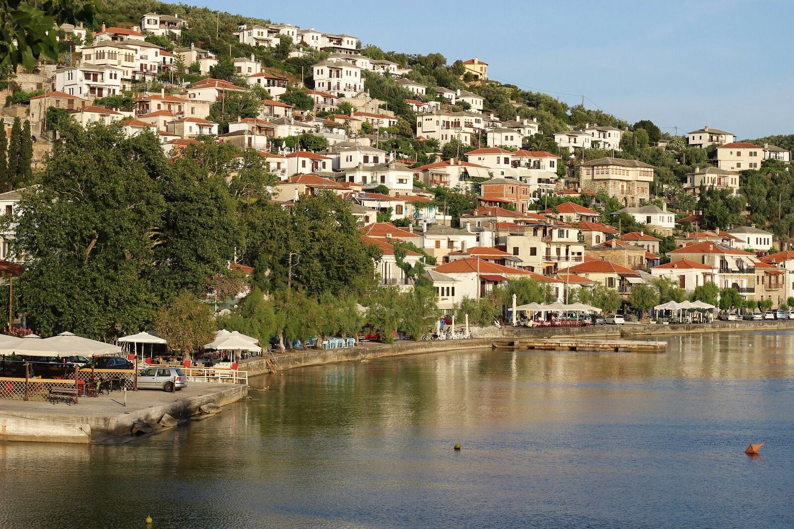 Afissos harbour on a June evening: white houses with tiled roofs climbing a green hillside, tavernas under plane trees along the quay, still water in front